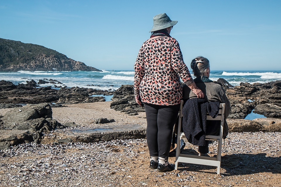 the back of two old ladies, one sitting one standing on a beach with sea in the distance and sand and rocks around them.