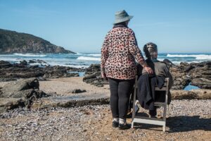 Two old ladies on a beach looking out to sea, one is sitting down, the other who is wearing a hat, is standing by her side
