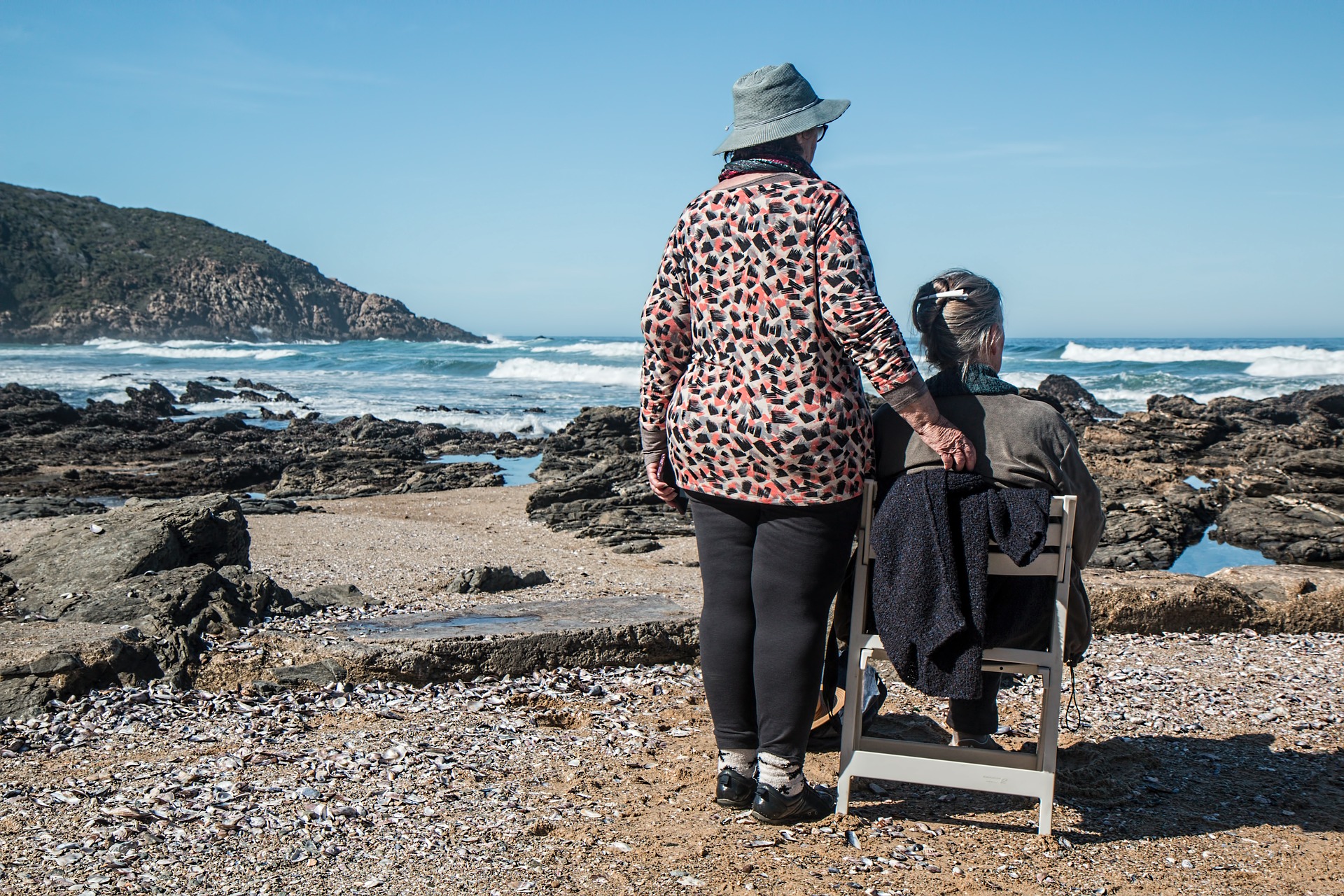 Two old ladies on a beach looking out to sea, one is sitting down, the other who is wearing a hat, is standing by her side